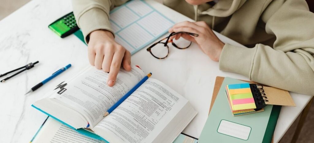 Estudar não é só ler. Livro aberto, pessoa acompanhando a leitura com o dedo. Muitos materiais de estudo na mesa.